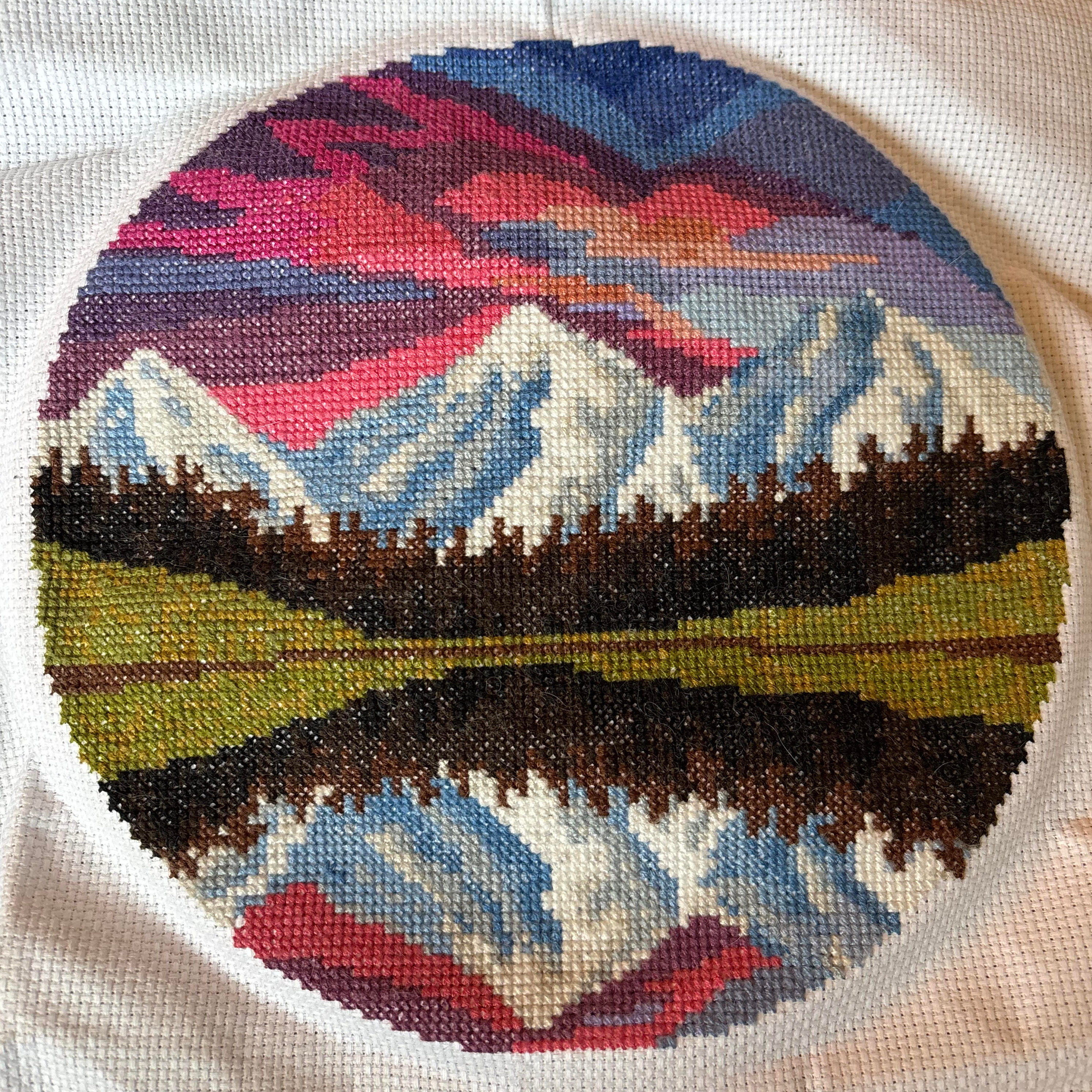 A circular image of snowy mountains overlooking a tree-lined reflecting lake. It is done in counted cross-stitch.